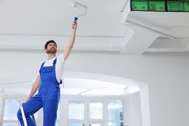 Photo of Handyman with roller painting ceiling on step ladder in room