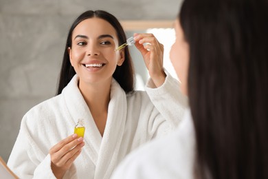 Beautiful young woman applying serum onto her face indoors