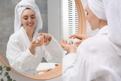 Photo of Beautiful woman in white robe applying cream near mirror indoors