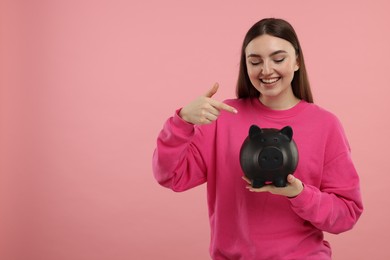 Happy woman pointing at piggy bank on pink background, space for text