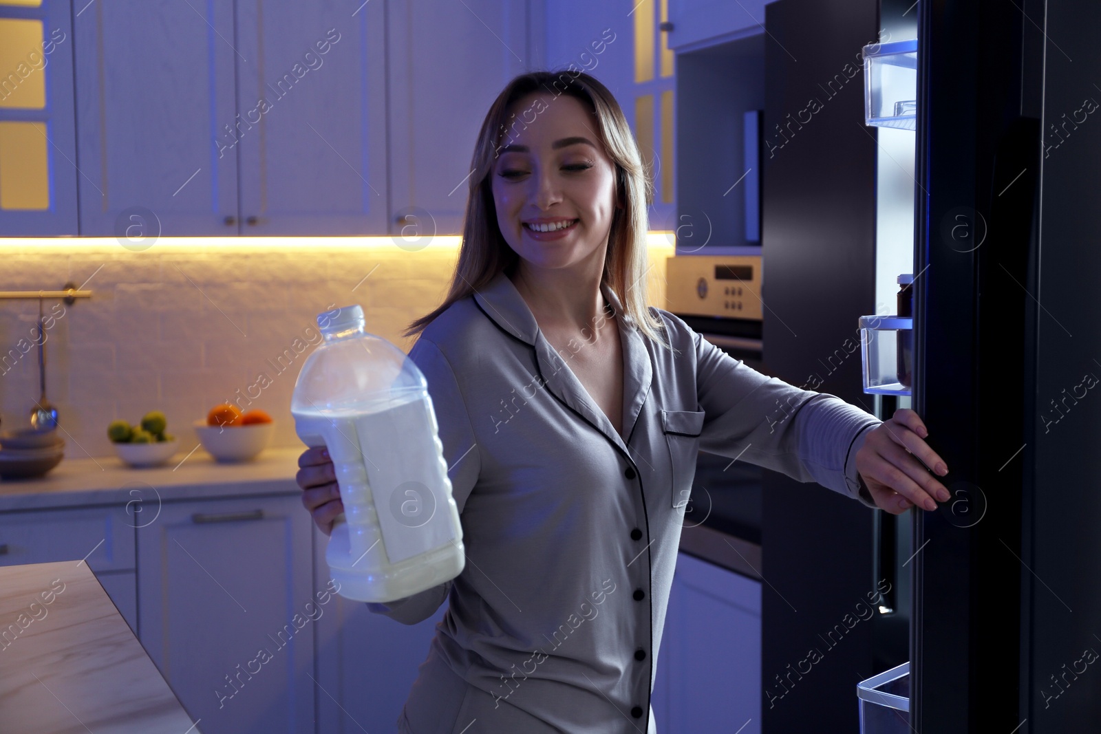 Photo of Young woman holding gallon bottle of milk near refrigerator in kitchen at night