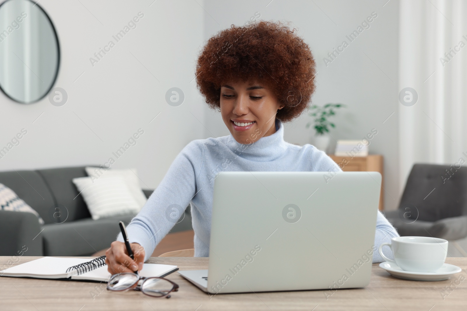 Photo of Beautiful young woman using laptop and writing in notebook at wooden desk in room