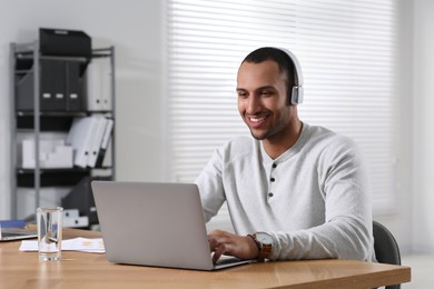 Photo of Young man with headphones working on laptop at table in office