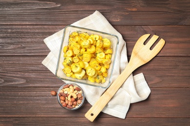 Baked yellow carrot in glass dish, nuts and fork on wooden table, flat lay