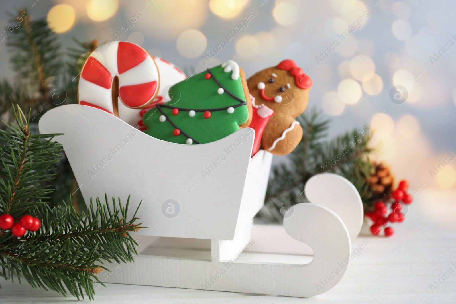 Image of Tasty Christmas cookies in decorative sleigh and fir branches on white wooden table against blurred fairy lights