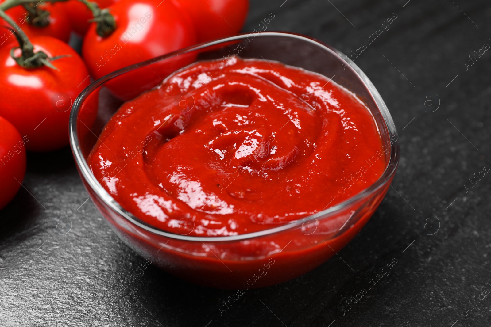 Photo of Organic ketchup in bowl and fresh tomatoes on black table, closeup. Tomato sauce