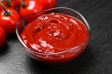 Photo of Organic ketchup in bowl and fresh tomatoes on black table, closeup. Tomato sauce