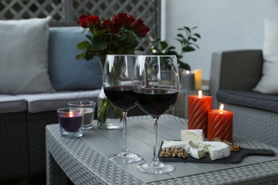 Glasses of wine, vase with roses, burning candles and snacks on rattan table at balcony in evening, closeup