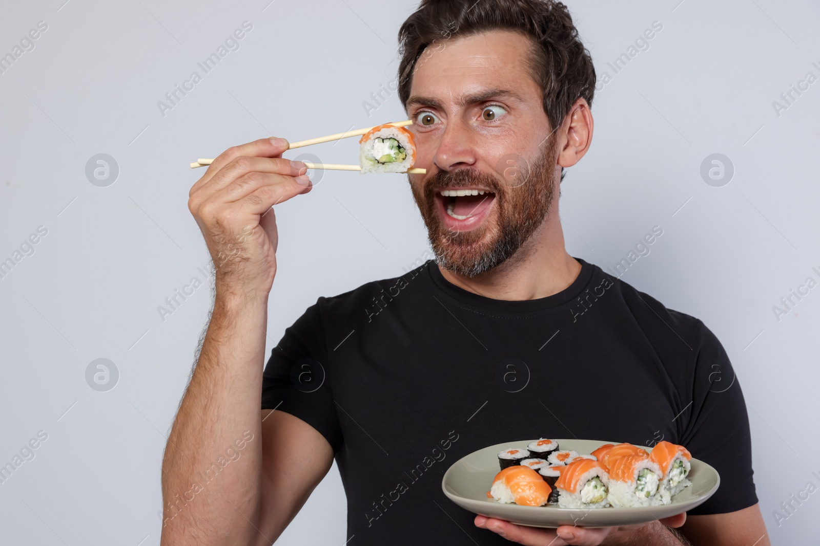 Photo of Emotional man eating tasty sushi roll and holding plate with food against light grey background, closeup