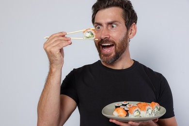 Photo of Emotional man eating tasty sushi roll and holding plate with food against light grey background, closeup