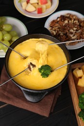 Dipping different products into fondue pot with melted cheese on black wooden table, above view