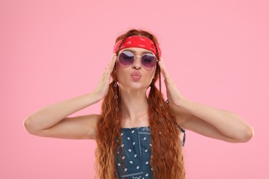 Photo of Stylish young hippie woman in sunglasses sending air kiss on pink background