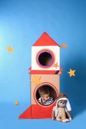 Photo of Cute little boy playing with cardboard rocket on light blue background