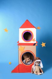 Cute little boy playing with cardboard rocket on light blue background