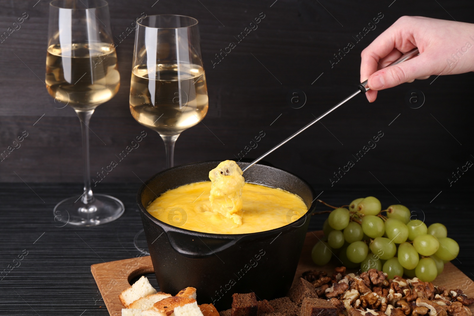 Photo of Woman dipping piece of bread into fondue pot with melted cheese at black wooden table, closeup