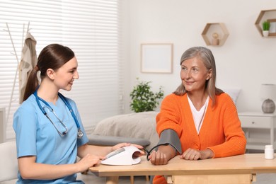 Young healthcare worker measuring senior woman's blood pressure at wooden table indoors