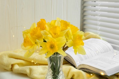 Photo of Beautiful yellow daffodils in vase, book and festive lights on windowsill