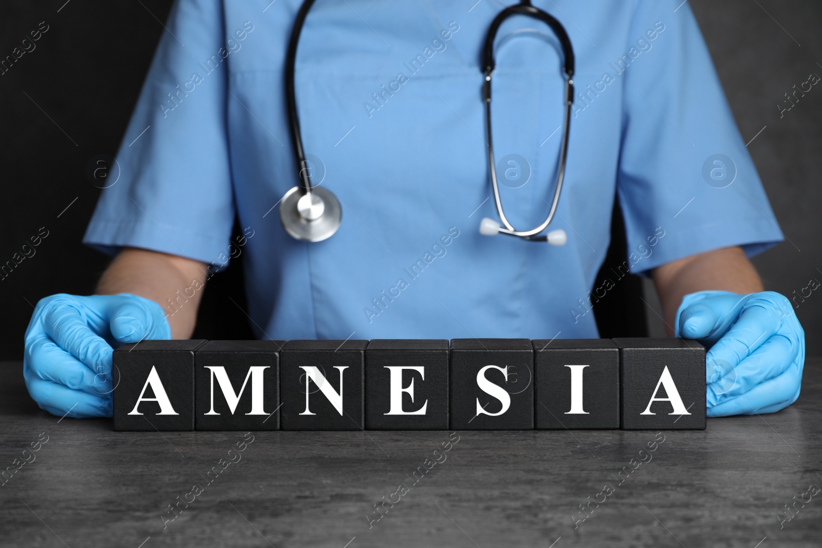 Photo of Doctor holding black cubes with word Amnesia at grey table, closeup