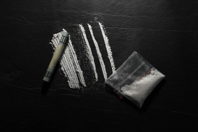 Photo of Drug addiction. Plastic bag with cocaine and rolled dollar banknote on dark textured background, flat lay