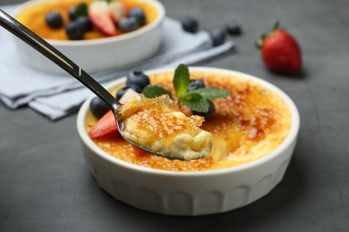 Photo of Eating delicious creme brulee with berries from spoon at grey table, closeup