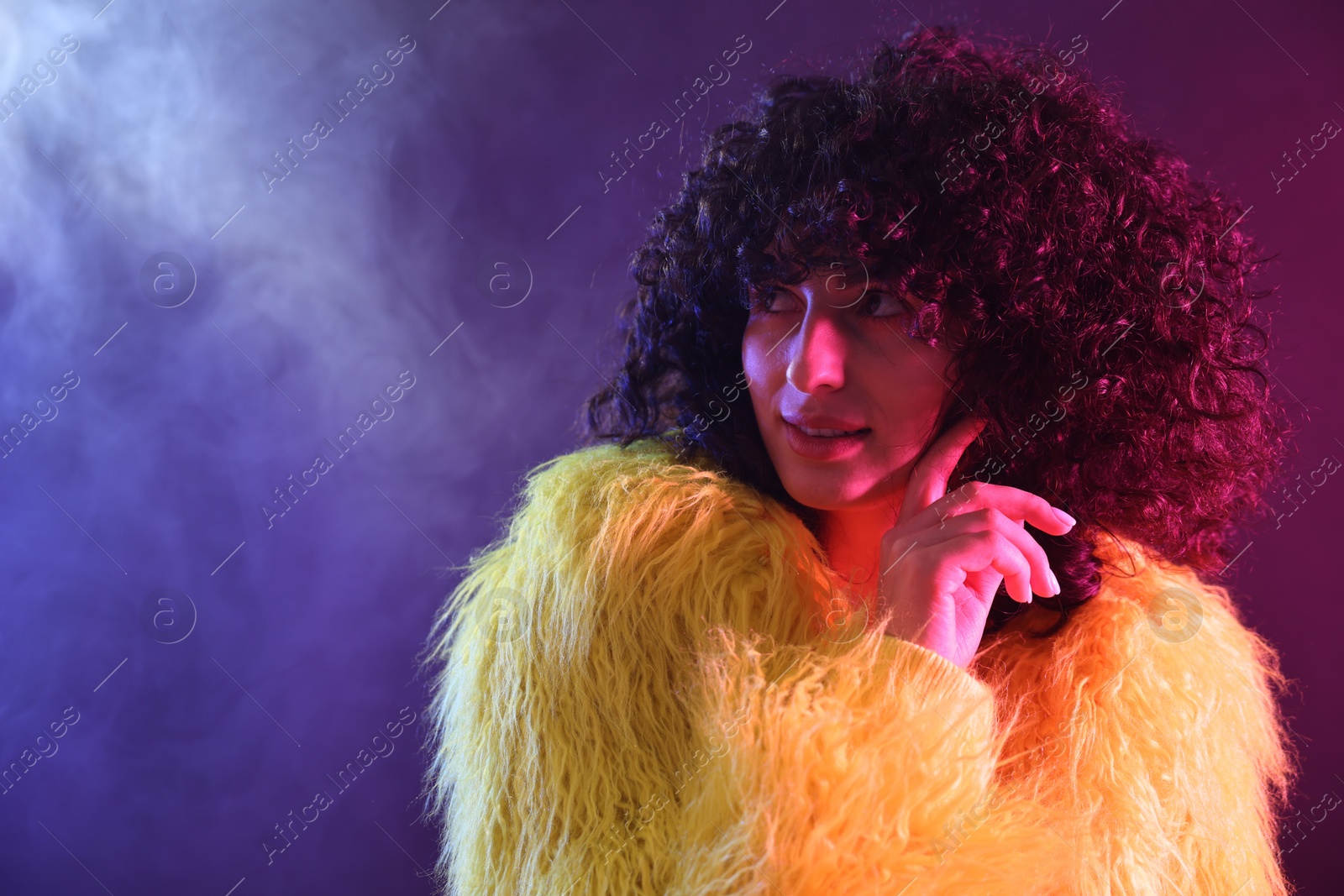 Photo of Beautiful young woman in yellow fur coat on color background in neon lights and smoke