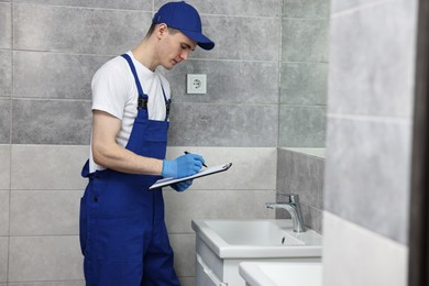 Photo of Young plumber writing results of examining faucet in bathroom