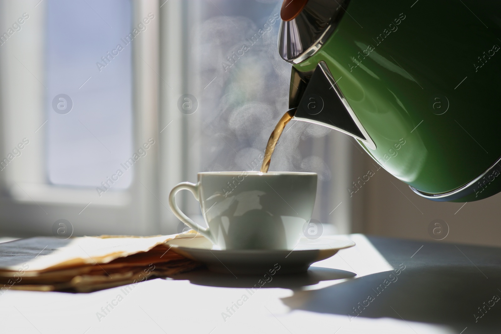 Photo of Pouring hot drink into cup near newspaper on sunlit table. Good morning