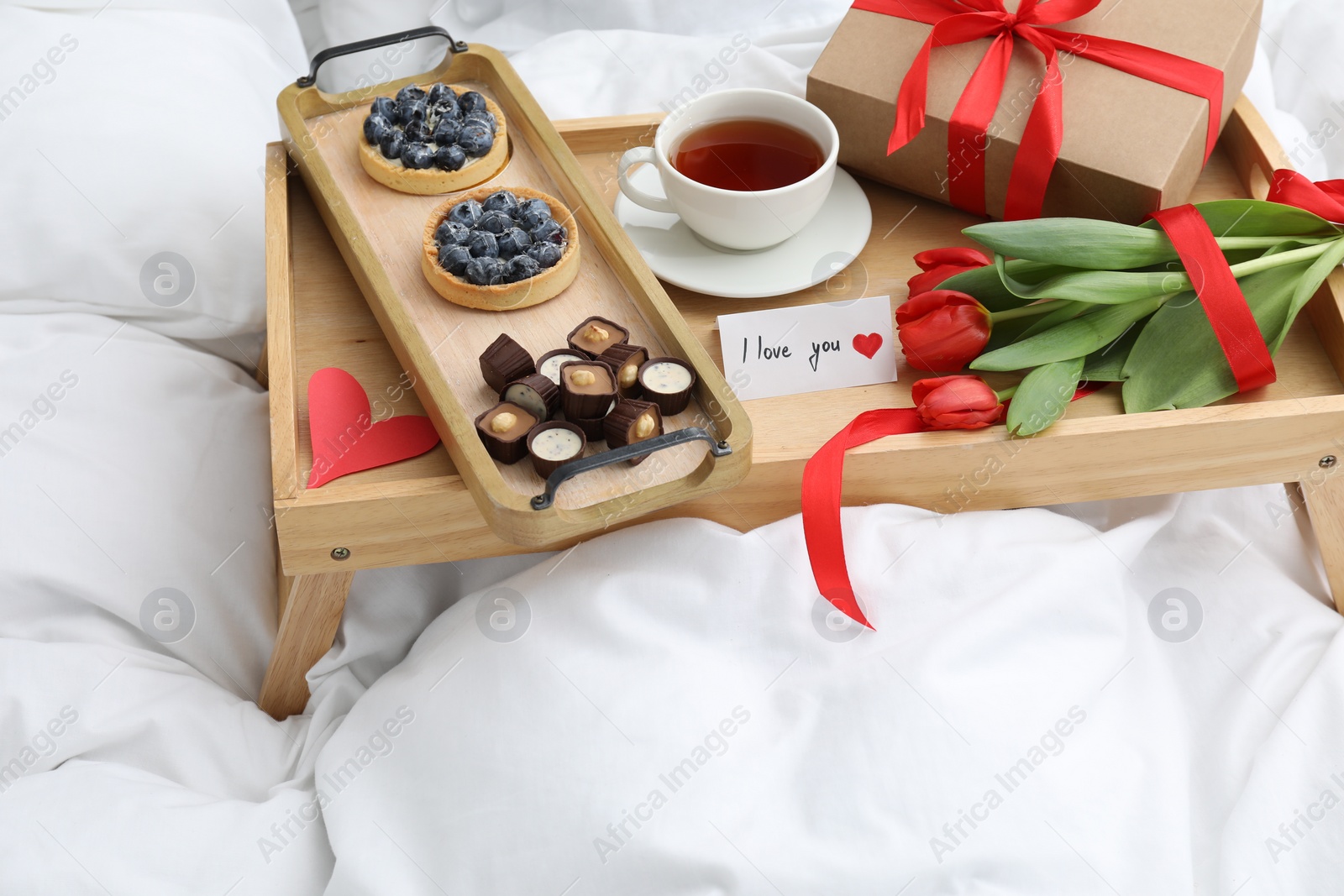 Photo of Tasty breakfast served in bed. Delicious desserts, tea, flowers, gift box and card with phrase I Love You on tray