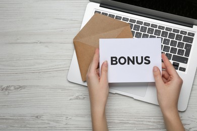 Image of Woman holding letter with word Bonus near laptop at white wooden table, top view