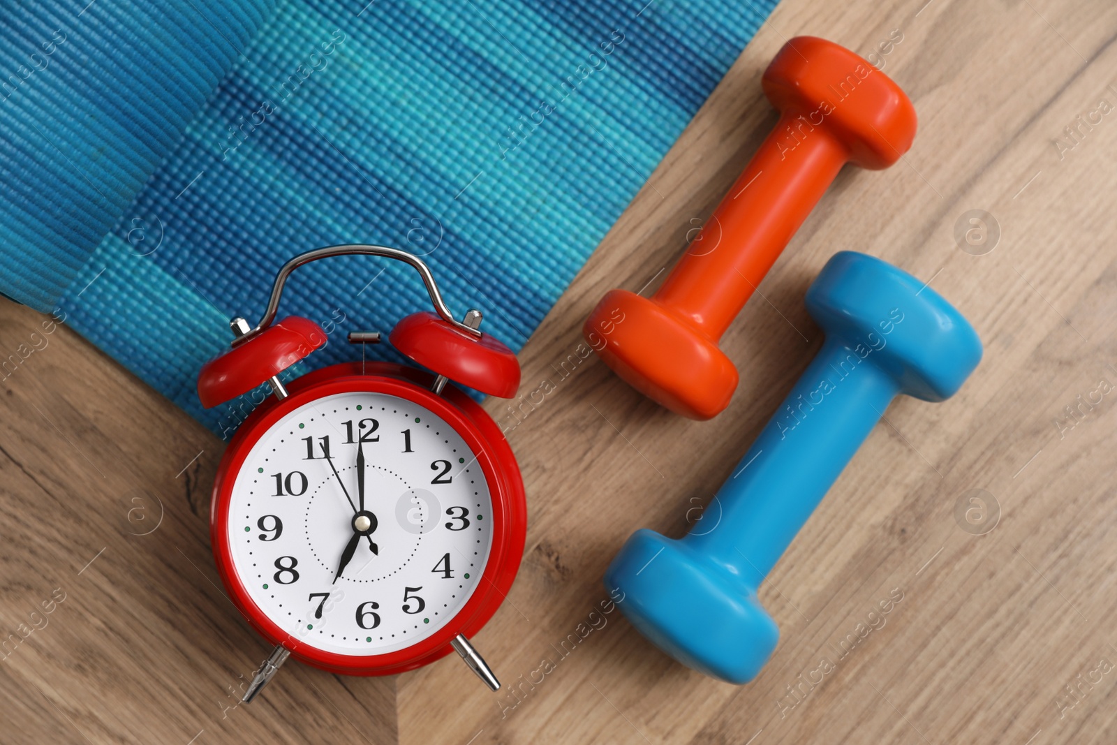 Photo of Alarm clock, yoga mat and dumbbells on wooden background, flat lay. Morning exercise