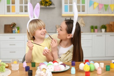 Mother and her cute son having fun while painting Easter eggs at table in kitchen