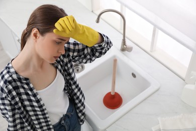 Photo of Tired young woman near clogged sink with plunger in kitchen, above view