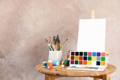 Photo of Wooden easel with blank canvas board and painting tools for children on table near color wall. Space for text