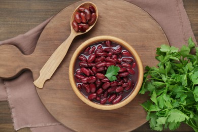 Photo of Bowl of canned red kidney beans, parsley and spoon on wooden table, flat lay