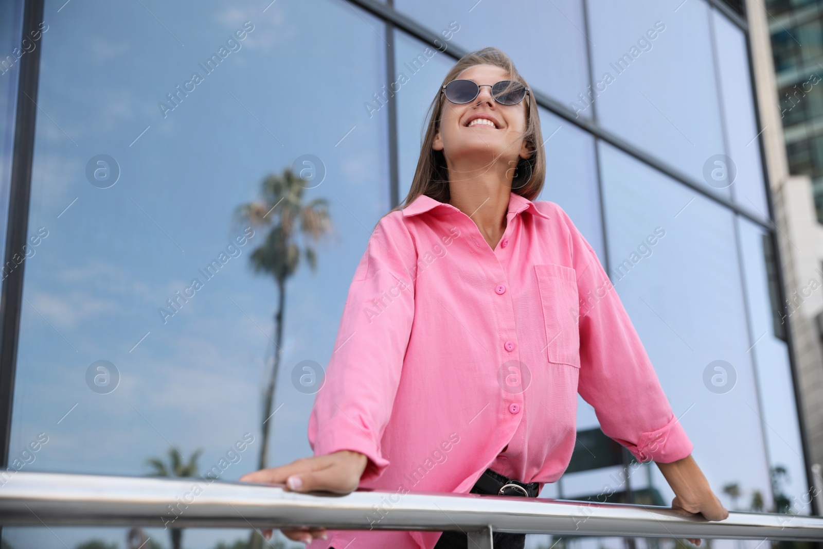 Photo of Beautiful young woman in stylish sunglasses holding onto railing near building outdoors