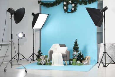 Photo of Beautiful Christmas themed photo zone with professional equipment, stylish armchair, trees and gift boxes in studio
