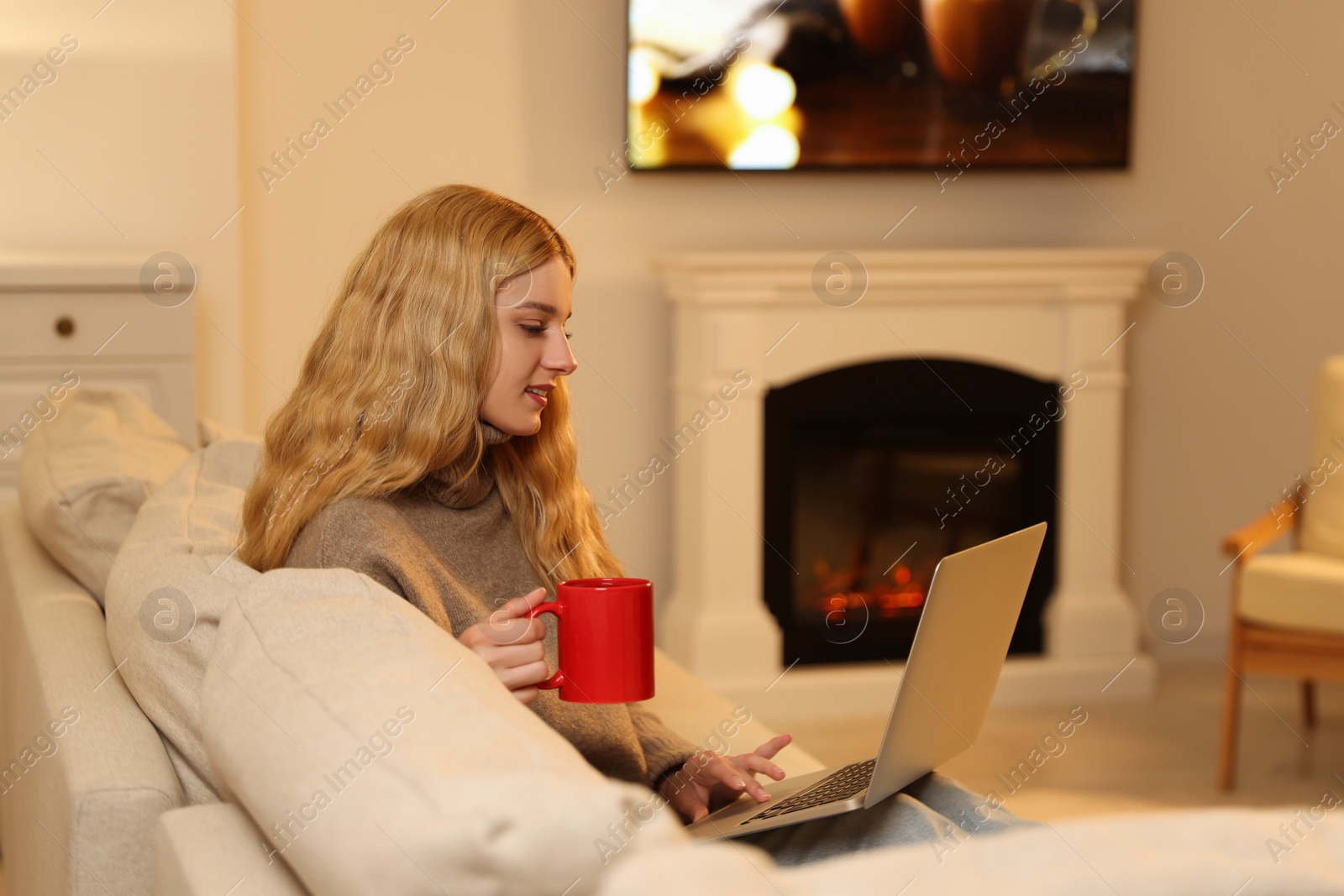 Photo of Young woman with cup of hot drink working on laptop near fireplace at home