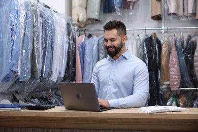 Photo of Dry-cleaning service. Happy worker using laptop at counter indoors