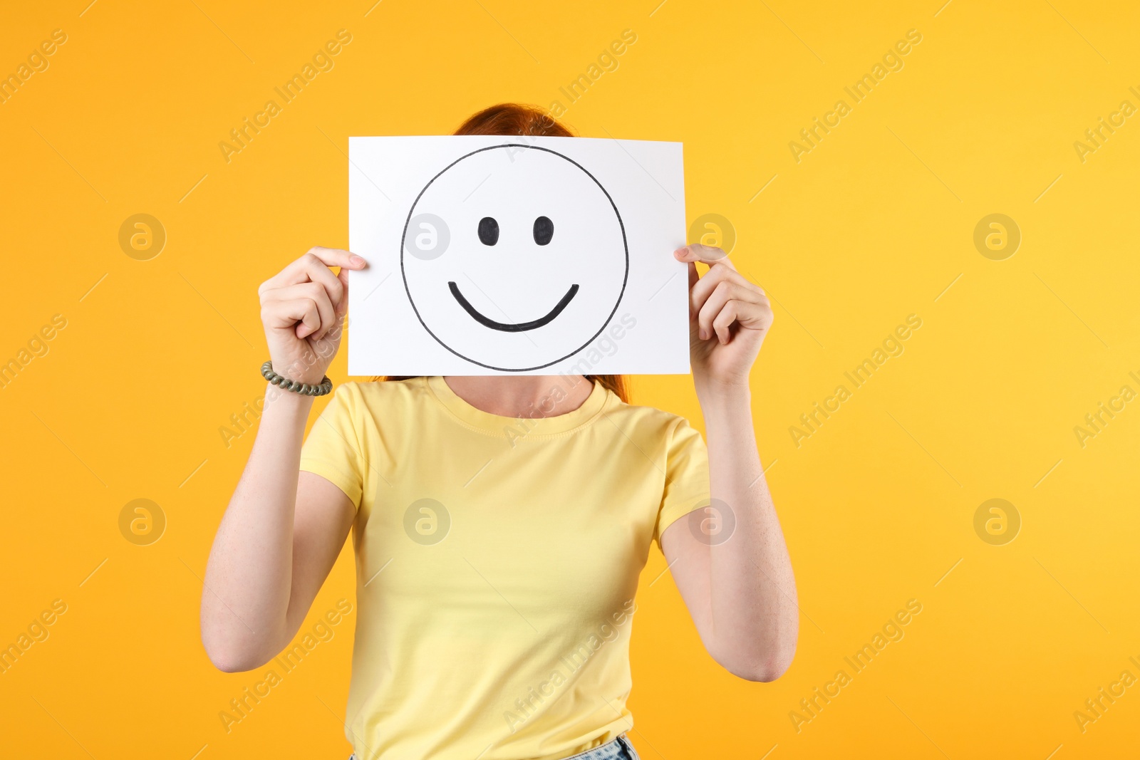 Photo of Woman hiding behind sheet of paper with happy face on yellow background