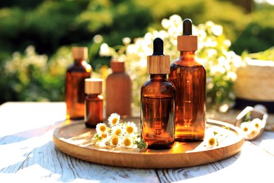 Bottles of essential oil and flowers on white wooden table outdoors, space for text