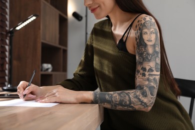 Beautiful woman with tattoos on arm drawing in sketchbook at table indoors, closeup