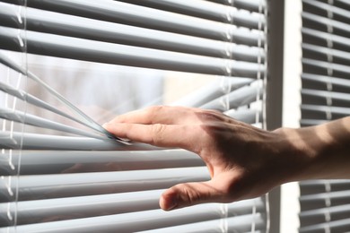 Man separating slats of white blinds indoors, closeup