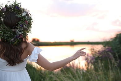 Photo of Young woman wearing wreath made of beautiful flowers outdoors at sunset, back view
