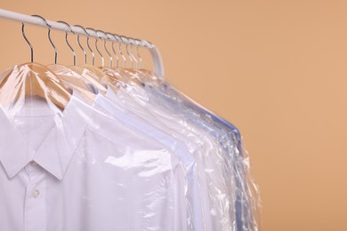 Photo of Dry-cleaning service. Many different clothes in plastic bags hanging on rack against beige background, closeup and space for text