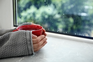 Photo of Woman with cup of hot tea near window on rainy day, closeup. Space for text