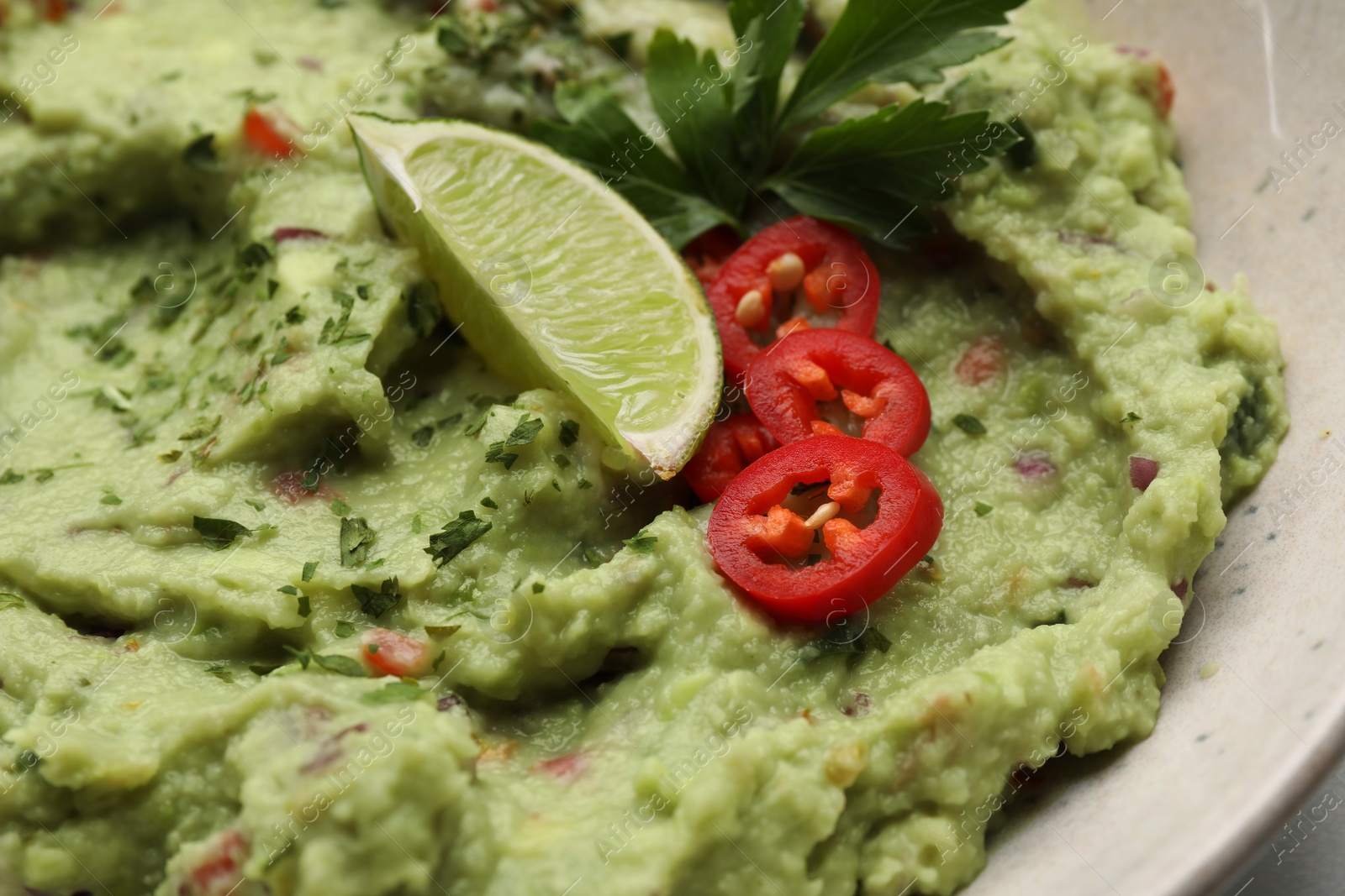 Photo of Bowl of delicious guacamole served with lime, pepper and parsley, closeup