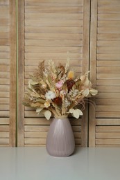 Photo of Beautiful dried flower bouquet in ceramic vase on white table near wooden folding screen