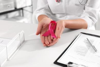 Photo of Doctor holding pink ribbon at white desk indoors, closeup. Breast cancer awareness