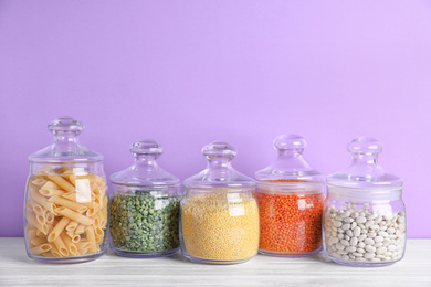 Photo of Jars with different cereals on white wooden table against violet background