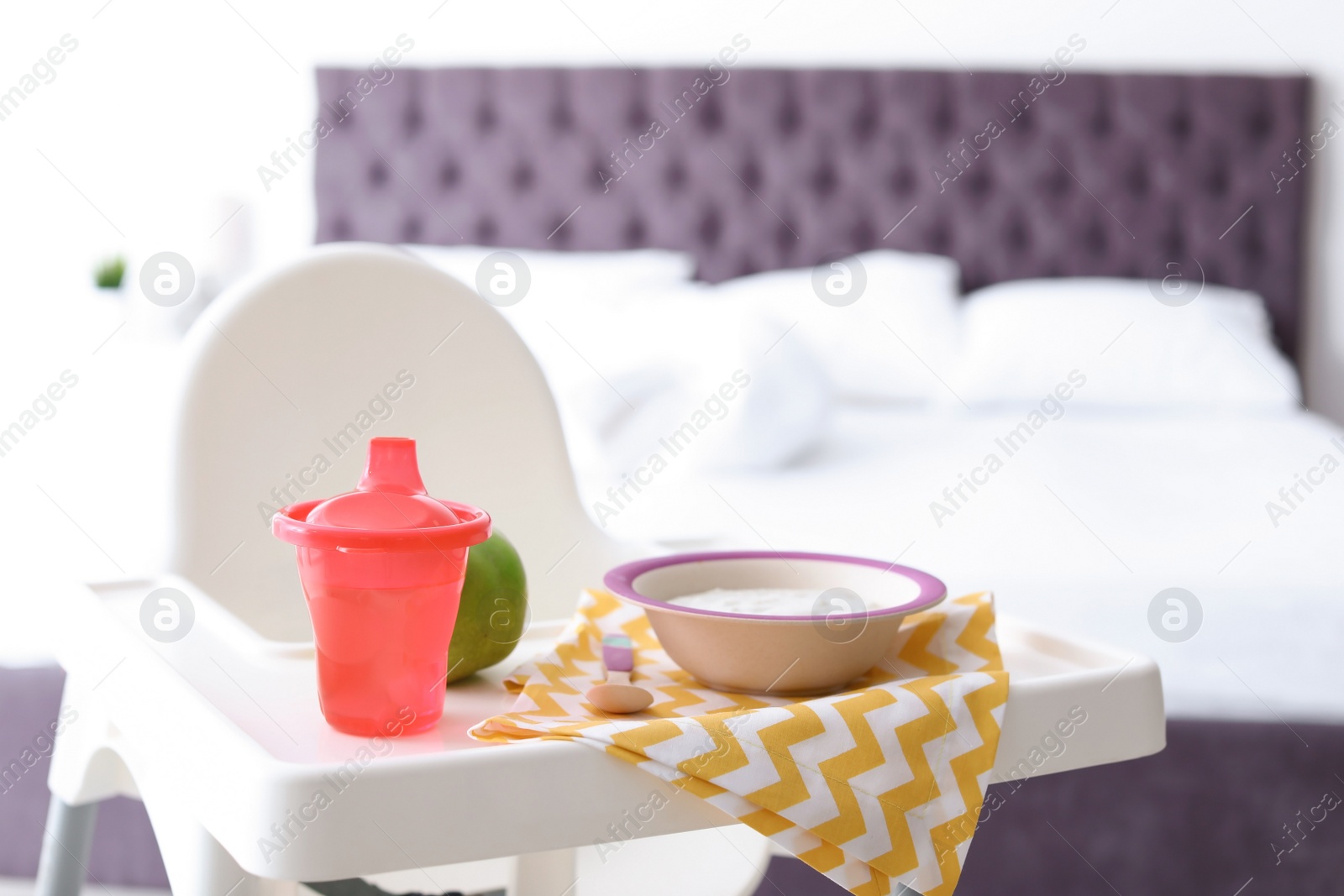 Photo of Bowl with delicious baby food, apple and drinking cup on highchair indoors
